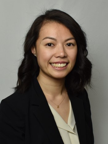 A woman with short dark wavy hair wears a cream-colored blouse, navy suit jacket, and gold necklace and smiles in front of a gray background.